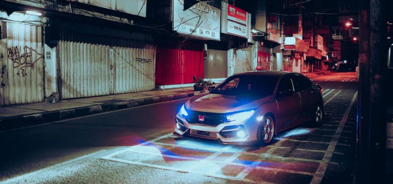 A car with headlights on in an empty urban street at night, surrounded by buildings.