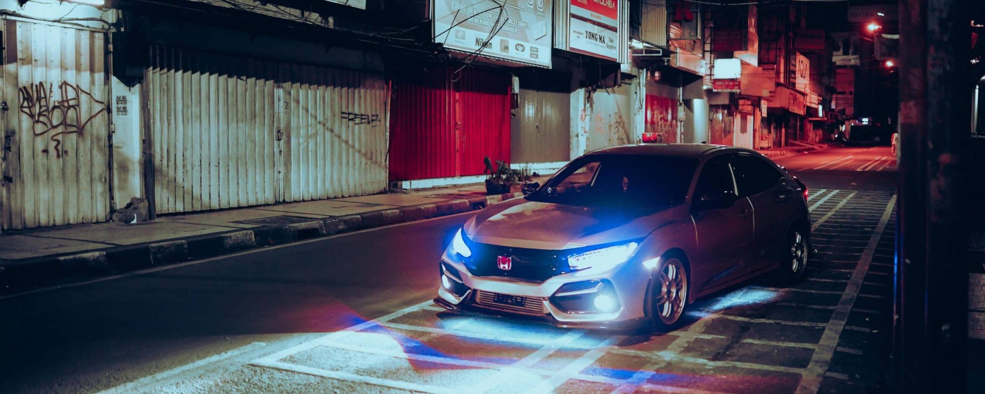 A car with headlights on in an empty urban street at night, surrounded by buildings.