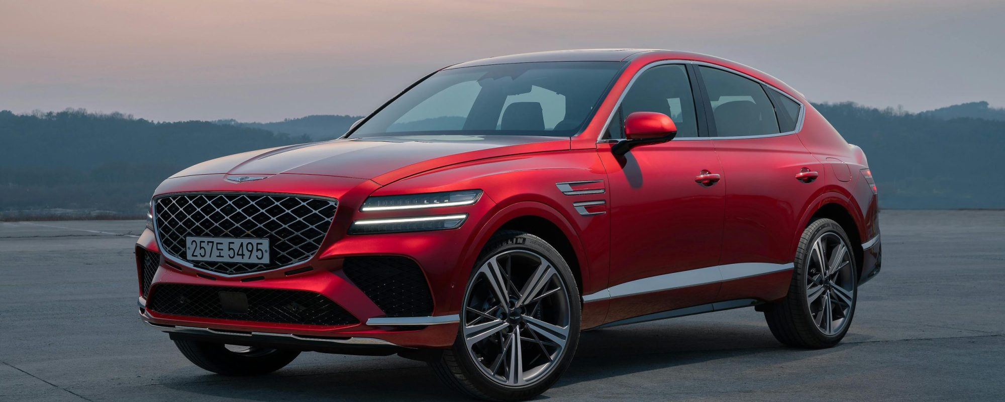A striking red Genesis GV80 SUV parked outdoors against a serene sunset backdrop.