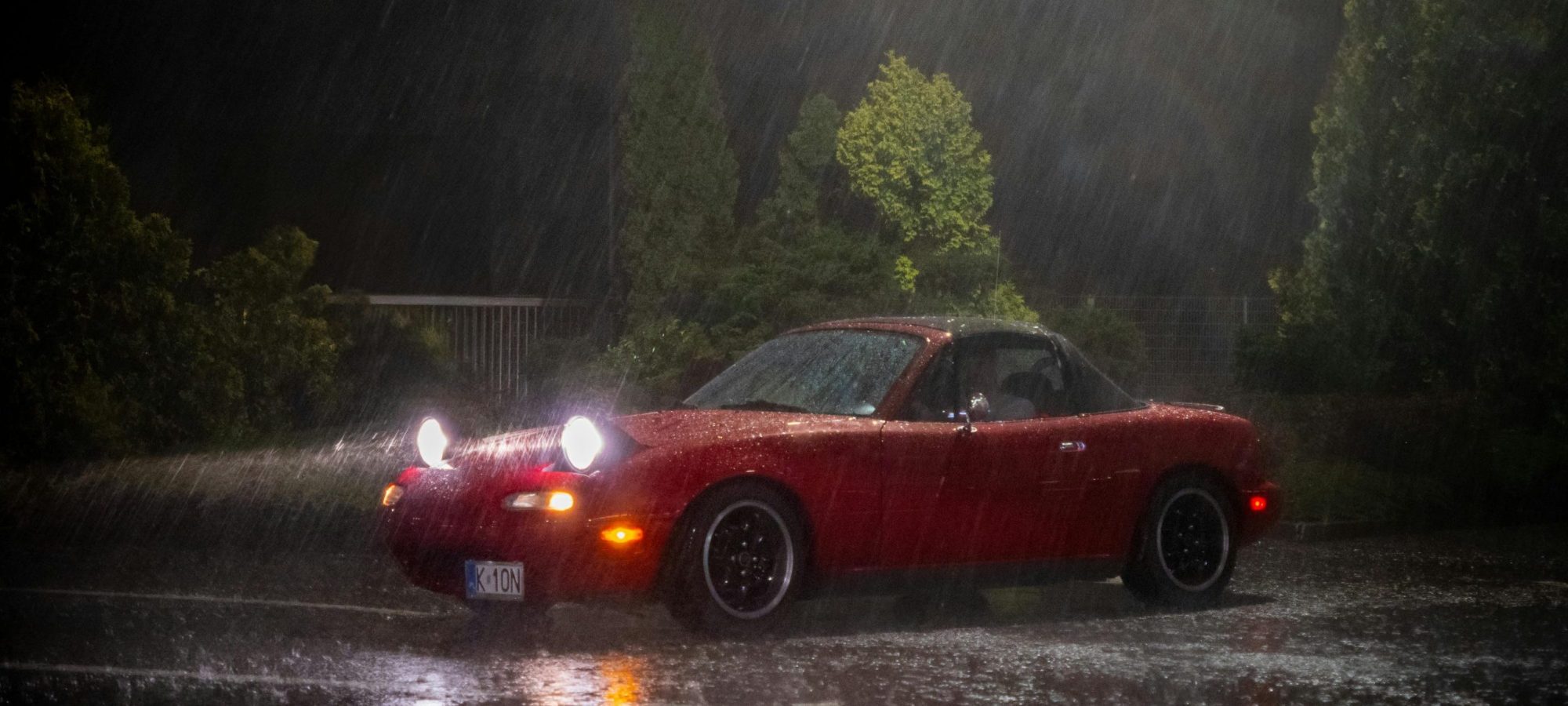 Red Mazda Miata parked in heavy rain at night, by Michal Robak. Maza Wheel Spacers / Adapters