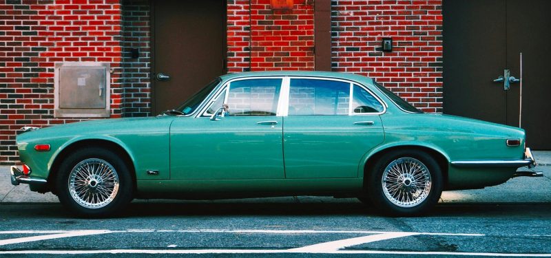 Vintage green Jaguar car parked in an urban setting against a red brick wall, highlighting classic automotive design.