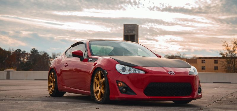 Stylish red sports car with golden rims parked outdoors under a cloudy sky.
