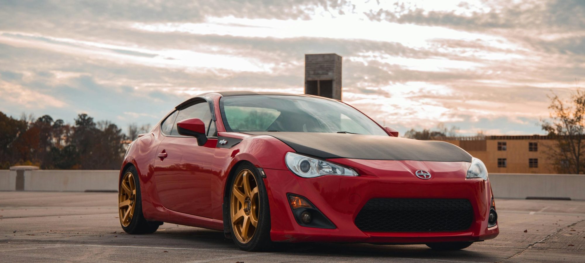 Stylish red sports car with golden rims parked outdoors under a cloudy sky.